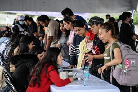 Latino people, some with children, give their details to women seated at a long table in a large tent