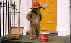 Paddington Bear, in his red hat, stands on the doorstep of his new home, hands on hips, looking down at a red bucket