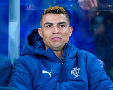 Cristiano Ronaldo sits on the substitutes bench in a blue padded jacket