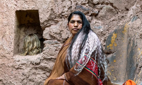 An older Indigenous woman with streaks of white hair and wearing a multicoloured shawl sits next to a blond wig in a carved aperture in a rock face