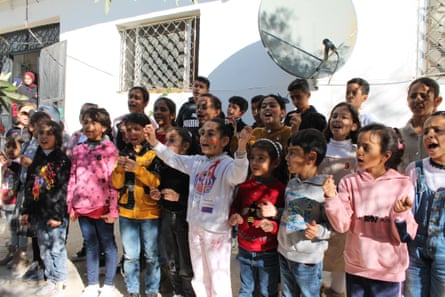 Children stand in rows outside a building, singing, some moving their hands or clapping in time to the music