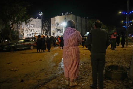 A woman in a bathrobe and other residents look on as Israeli security forces and rescue teams work at the damaged site