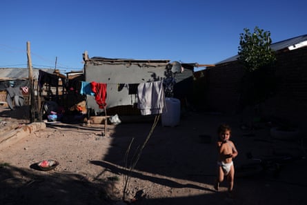 A child in a nappy runs in a dirt yard behind a house made of tarpaulin and tin