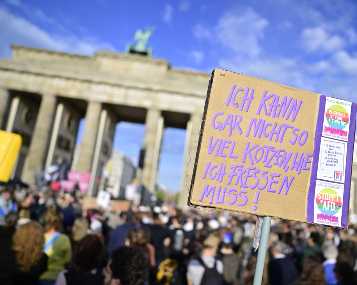 A protest sign with purple handwritten German text stands above a crowd near the Brandenburg Gate