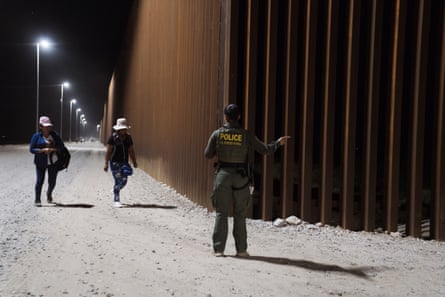 a law enforcement officer points in a direction along a border fence as two people walk near her