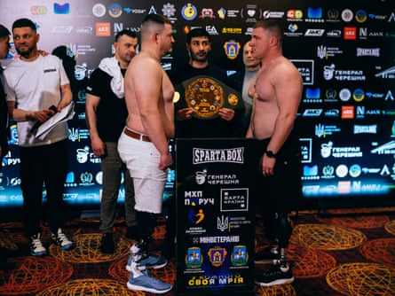 Khrebet and Drobotenko facing each other either side of a podium. A man stands between them holding the belt