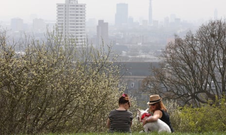 Two women and a dog sit on Parliament Hill in Hampstead Heath overlooking an overcast Central London
