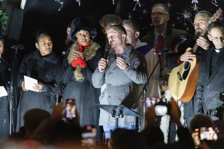 Tommy Robinson speaks into a microphone on a stage. He is standing next to a man holding an acoustic guitar and there are women beside him who look as if they are preparing to sing. A crowd is around him.