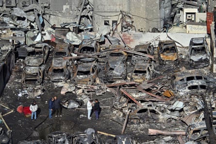 Burnt shells of about a dozen cars amid rubble as several people look on.