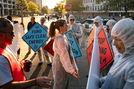 A group of people wearing white mesh suits carry signs outside with messages such as ‘Fund safe clean energy’