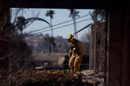 Under a clear sky with palm trees in the background, a man in a yellow uniform and helmet looks down at rubble.