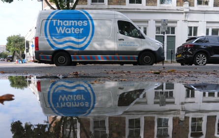 A white van with the blue Thames Water logo is parked next to a large puddle or flooded area on a street in front of white houses. The van and logo are reflected in the water.