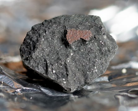 A broken piece of black and grey rock studded with white flecks with a small reddish area, sitting on tin foil.