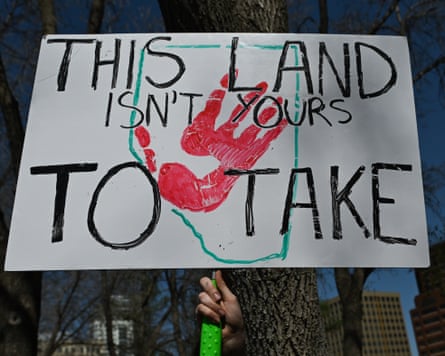 A protest sign reading “This Land Isn’t Yours To Take” with a red handprint, held outdoors in Edmonton