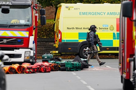 An armed officer walks past an ambulance near fire engines, with emergency services equipment on the ground