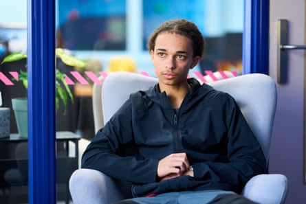Remy sits in a pale blue armchair in front of a glass wall with bright blue frames. He is small and thin with his hair tied back and wears a black zip-up hooded anorak.