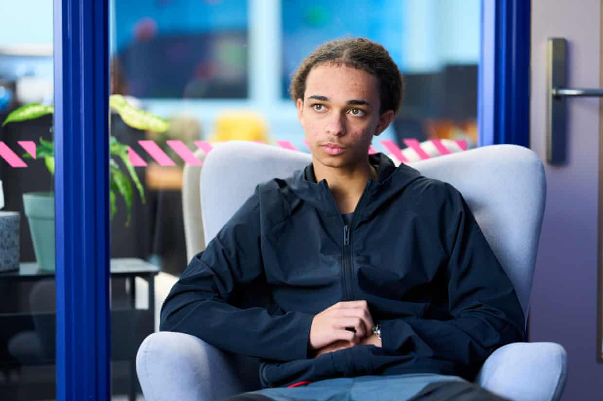 Remy sits in a pale blue armchair in front of a glass wall with bright blue frames. He is small and thin with his hair tied back and wears a black zip-up hooded anorak.