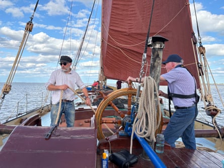 Two men on the deck of a red-masted Thames sailing barge. One holds the wheel while the other is pulling a rope.