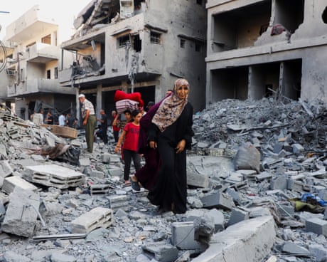 Aftermath of an overnight Israeli strike on a house in Gaza City: a woman and children pick their way through rubble in front of destroyed buildings. One boy is holding a pile of bedding on his head; men are standing in the background surveying damage.
