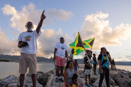 Wilbourn Carr points upward on rocks by the sea, surrounded by campaigners and a Jamaican flag