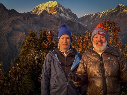 Two men in puffa jackets and woolly hats pose in front of a spectacular mountain peak