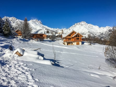 A snowy slope with a few wooden chalets on it and mountain peaks behind
