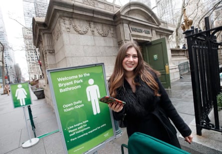 woman stands next to sign that says ‘welcome to the bryant park bathroom’, with a fancy Beaux-Arts building behind her saying ‘women- men’
