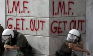 Greek riot policemen resting in front of graffiti reading 'IMF get out'