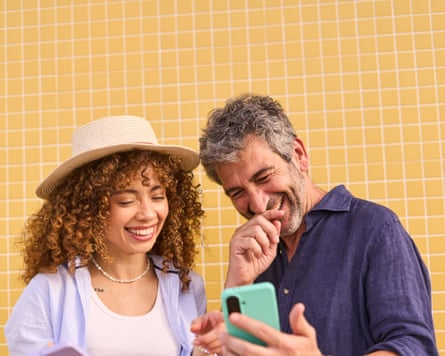 A couple laughing using a smartphone as they stand in front of a yellow tiled wall.