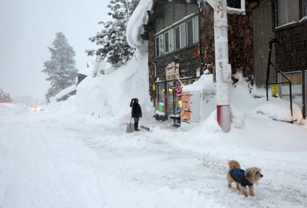 A woman and her dog stand in the snow in front of a wood front store in a mountain town.