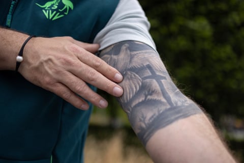 A man’s hand to left touches his upper arm where a cross and a white dove are tattoo on his skin