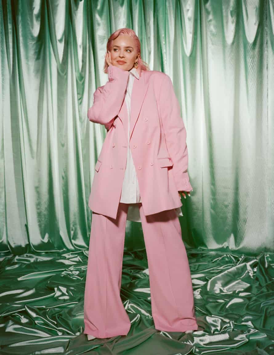 Anne-Marie in an oversized pink trouser suit and white blouse with masses of green satin on the floor and as a curtain behind her