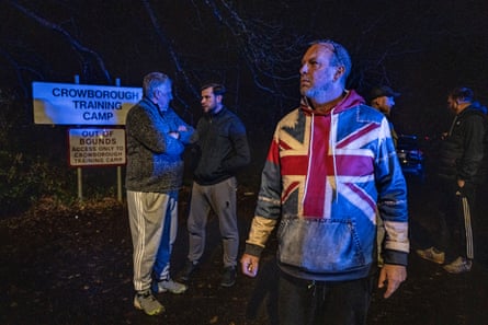 A sombre-looking man in a union jack denim hoodie among men standing beside the sign for Crowborough training centre