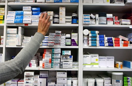 A worker in a pharmacy reaches up to pick out a box of medication from a high shelf