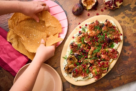 A platter of labneh, figs, pomegranate and herbs on a wooden table. To the sides, a pair of hands break up a crispy pita.