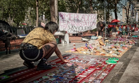 A wona crouches  on the ground to arrange products on her stall. Behind her is a sign in Spanish which reads ‘Feminist Market against economic violence’.