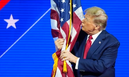 Older man in dark suit has arms around standing US flag and is pursing his lips.