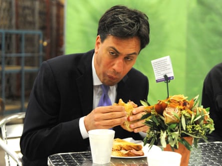 Ed Miliband sitting at a silver metal table on a metal chair, wearing a suit and tie, and making a very unfortunate face while trying to eat a bacon sandwich