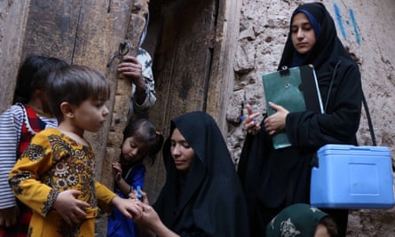 Two women in hijabs, one with a coolbox over her shoulder and a clipboard, stand at a door with small children