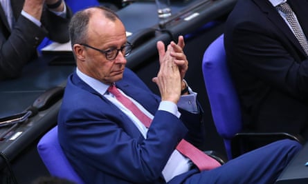 Friedrich Merz, in a suit and tie and wearing glasses, frowns as he holds his hands up to clap