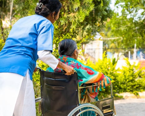 An elderly woman in a wheelchair being pushed by a carer through a tropical garden