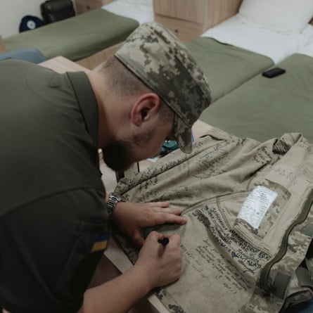 A male cadet in a camouflage cap writes something in black marker pen on the inside of a camouflage shirt, where there are other messages.