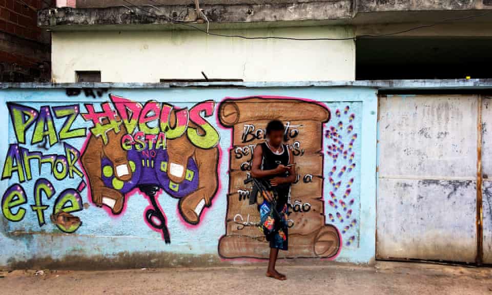 A heavily armed drug trafficker stands near a mural that reads ‘God's in control’ in one Rio favela