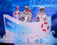 Four people wearing sunglasses and shiny silver outfits behind a piece of stage scenery representing a boat