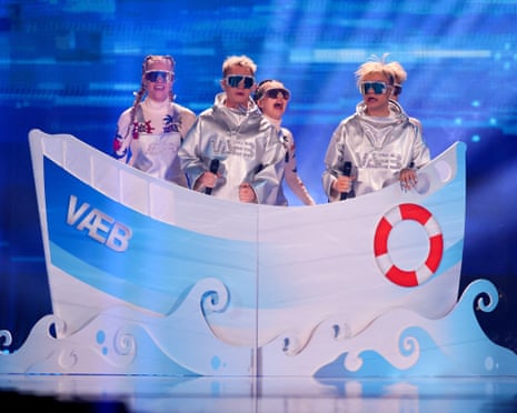 Four people wearing sunglasses and shiny silver outfits behind a piece of stage scenery representing a boat