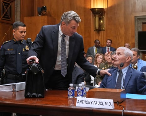 David Schertler stands and grasps Anthony Fauci by the shoulder as a police officer waits to escort him out