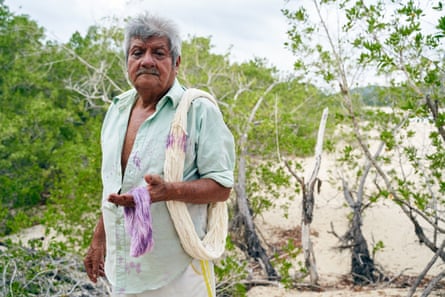 A older man stands on a beach holding white yarn, stained purple in places