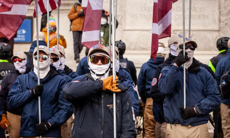 Members of the white supremacist group Patriot Front, dressed in khakis, navy blue jackets, facemask, sunglasses and khaki baseball caps, hold flags as they stand in formation.