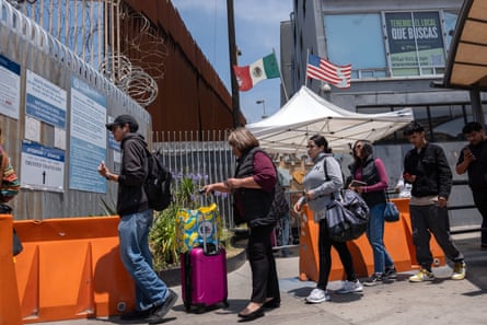 People line up at a gate with Mexican and American flags in the background.