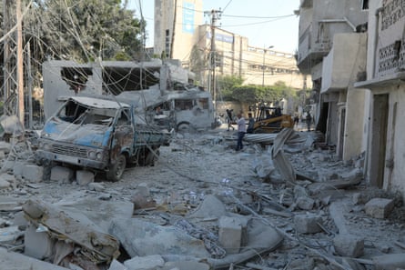 Men try to clear fallen power cables and debris in a street with ruined trucks and vehicles. Rubble, mattresses and household items are strewn around under a layer of thick grey dust.